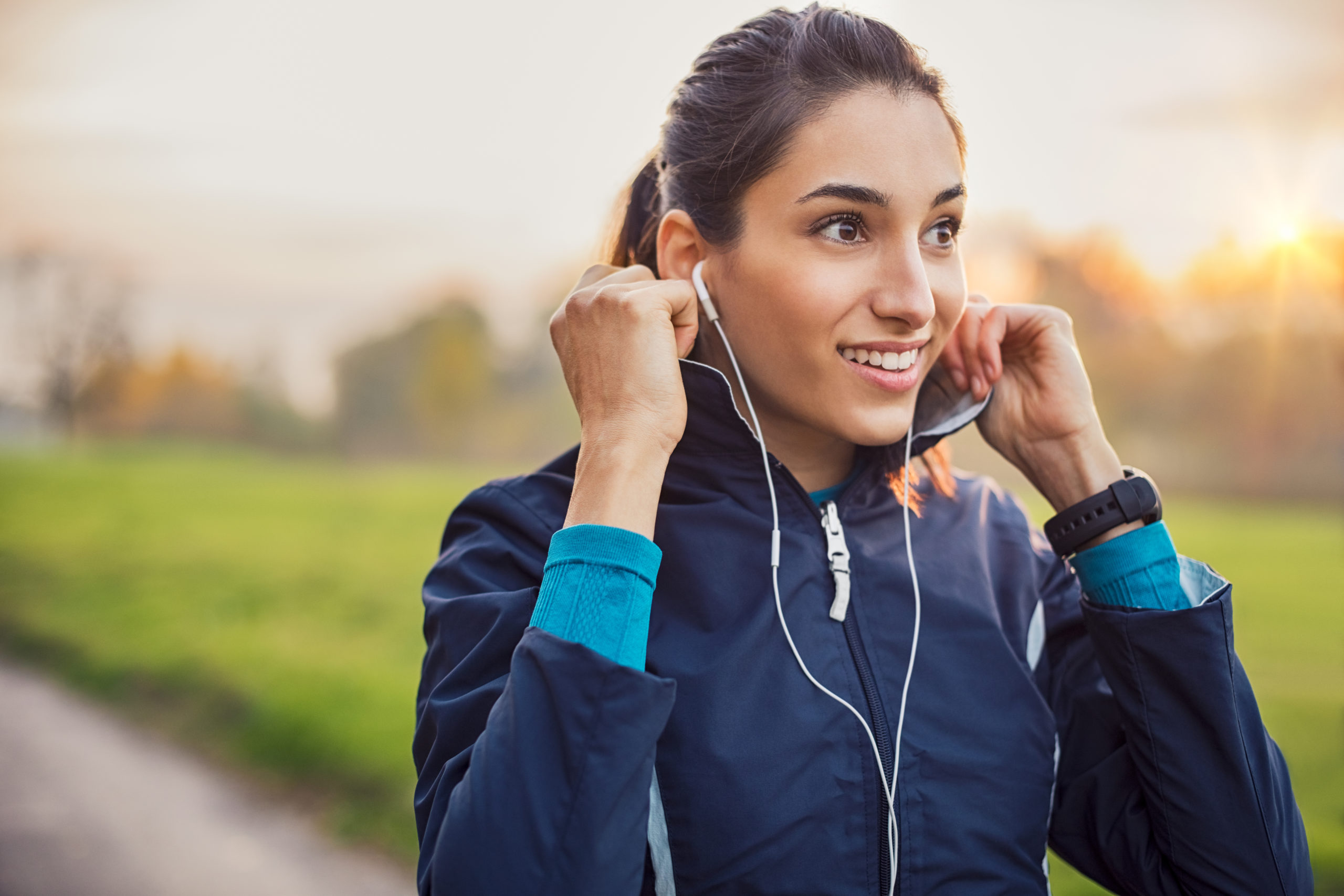 Young athlete adjusting jacket while listening to music at park. Smiling young woman feeling relaxed after a long run during the sunset. Happy sporty woman smiling and looking away during workout.
