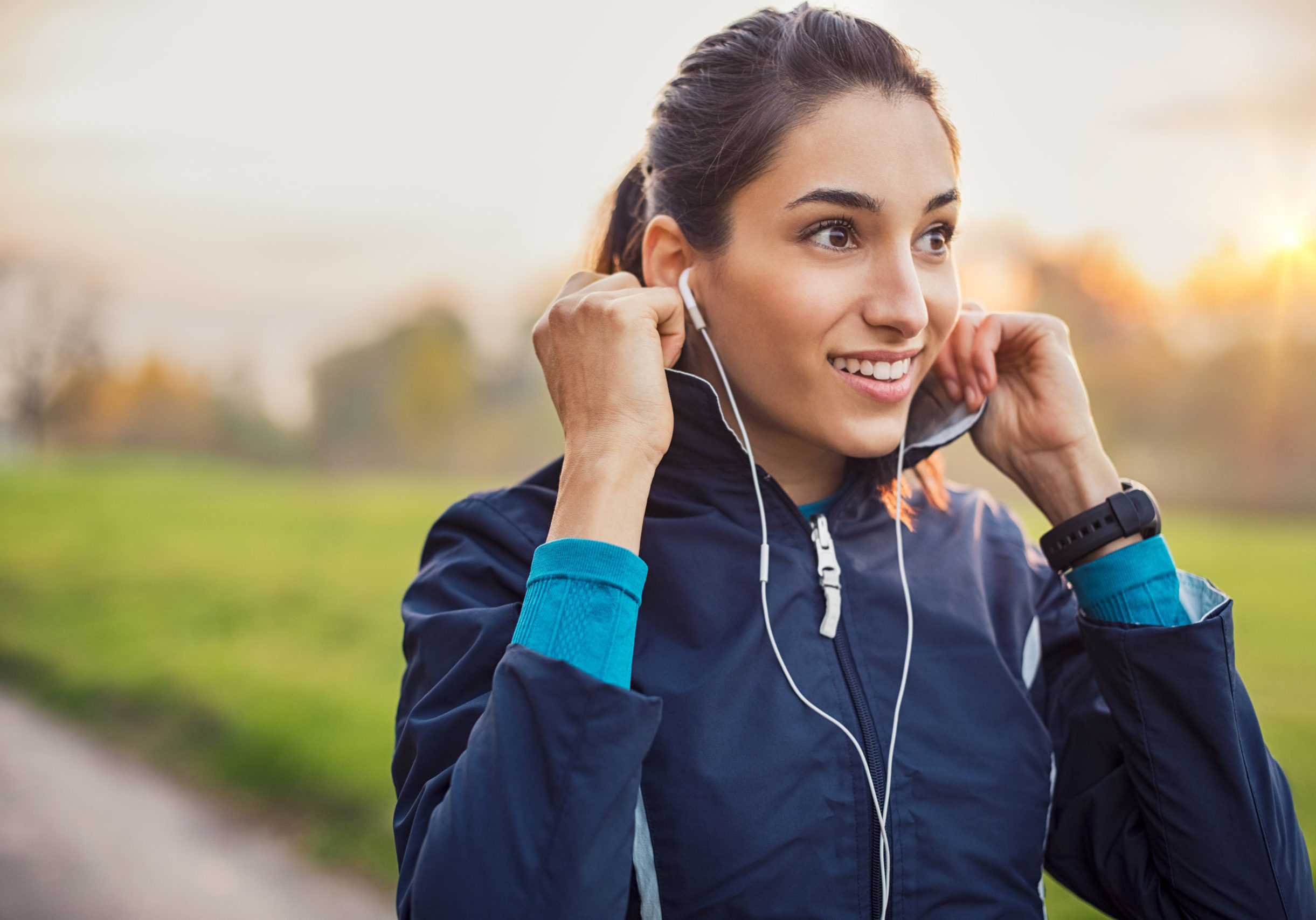 Young athlete adjusting jacket while listening to music at park. Smiling young woman feeling relaxed after a long run during the sunset. Happy sporty woman smiling and looking away during workout.